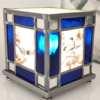 Lampe vitrail Azulero en forme de cube. Dessins d'oiseaux sur les quatre faces et bordure en verre bleu et miroir. Lampe présentée de trois quart, allumée