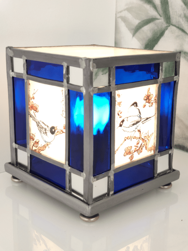 Lampe vitrail Azulero en forme de cube. Dessins d'oiseaux sur les quatre faces et bordure en verre bleu et miroir. Lampe présentée de trois quart, allumée
