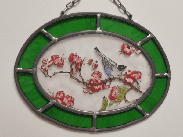 médaillon Cestya de forme ovale avec chainette d'accroche et un liseré vert. Il représente un petit oiseau sur une branche de cerisier