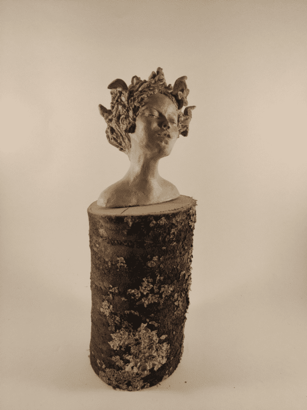 Tête de femme posée sur un tronc de bois. Elle ferme les yeux et ses cheveux avec des fleurs s'envolent au vent. pièce terre crue beige cirée. Vue de trois quart