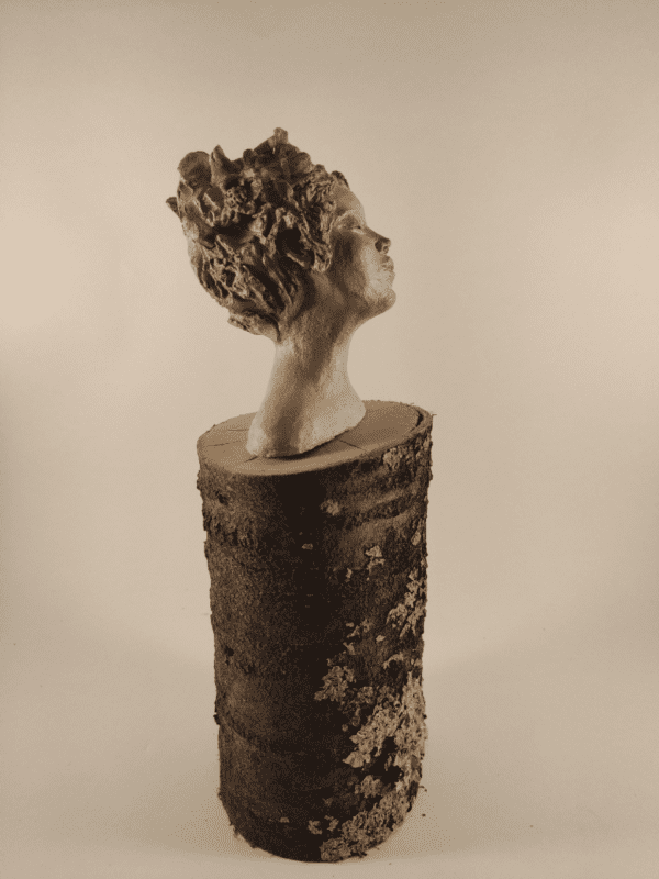 Tête de femme posée sur un tronc de bois. Elle ferme les yeux et ses cheveux avec des fleurs s'envolent au vent. pièce terre crue beige cirée. Vue de profil