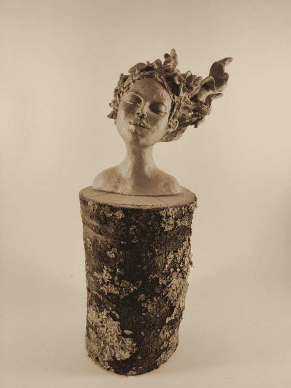 Tête de femme posée sur un tronc de bois. Elle ferme les yeux et ses cheveux avec des fleurs s'envolent au vent. pièce terre crue beige cirée. Vue de trois quart