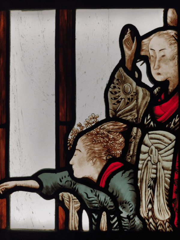 vitrail inséré dans une boite lumineuse représentant deux Geishas derrière une fenêtre en bois. Elles observent quelque chose au loin. Un des personnages montre quelque chose du doigt. le vitrail s'intitule "le retour"