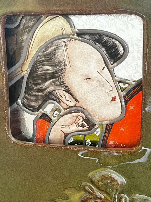 Vitrail représentant le portrait d'une geisha. Il est inséré dans un cadre en céramique émaillé vert avec des roses en relief. Vue de près. Elle s'intitule Mouna