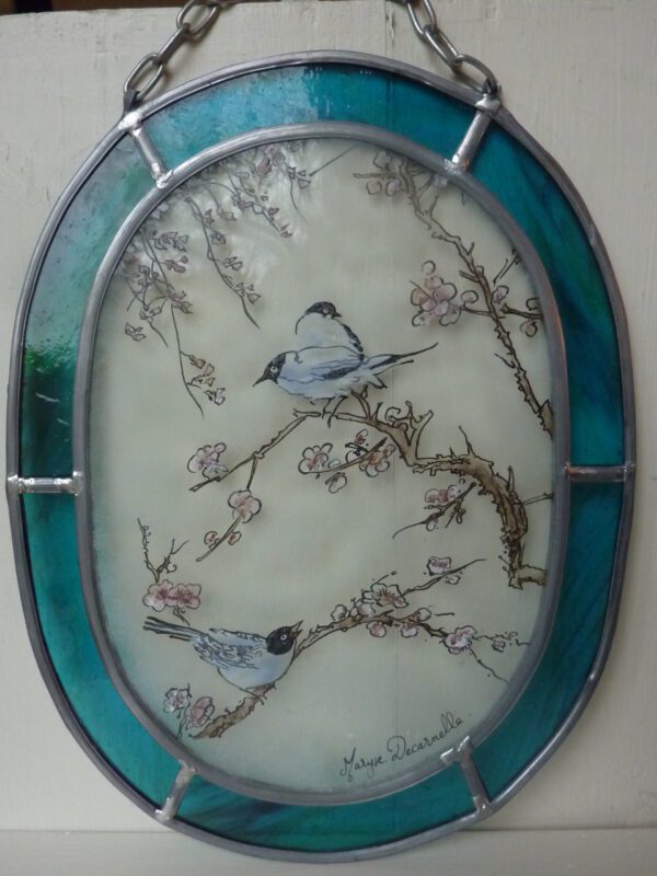 médaillon ovale avec une chainette permettant de l'accrocher. Il à un liseré bleu et son centre est décoré avec trois mésanges sur des branches de cerisiers en fleur. Vue de près. Il s'intitule Pitouine