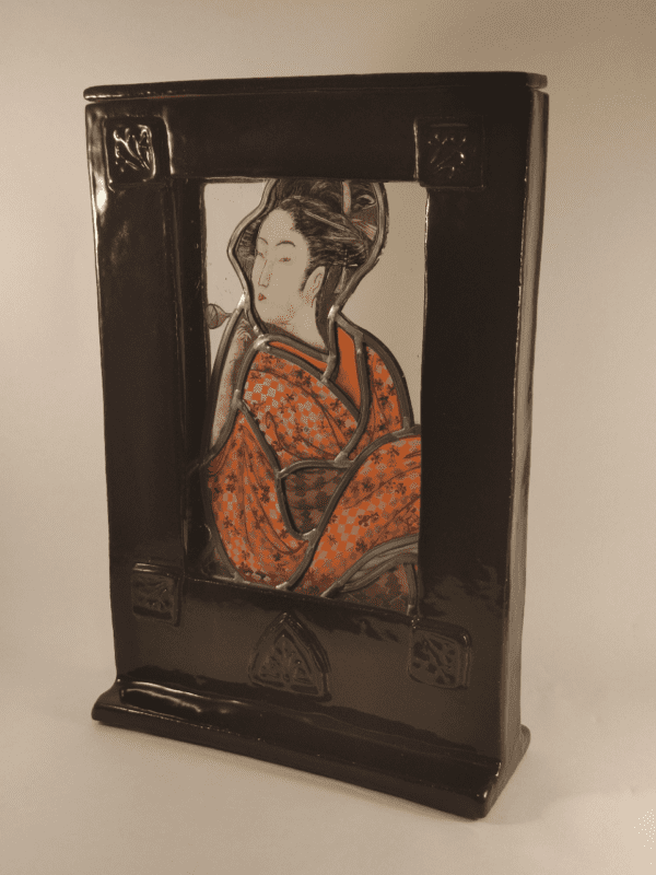 Cadre vitrail Terra. Un vitrail avec une geisha en kimono orange est inséré dans un cadre en céramique émaillé noire. Vue de trois quart