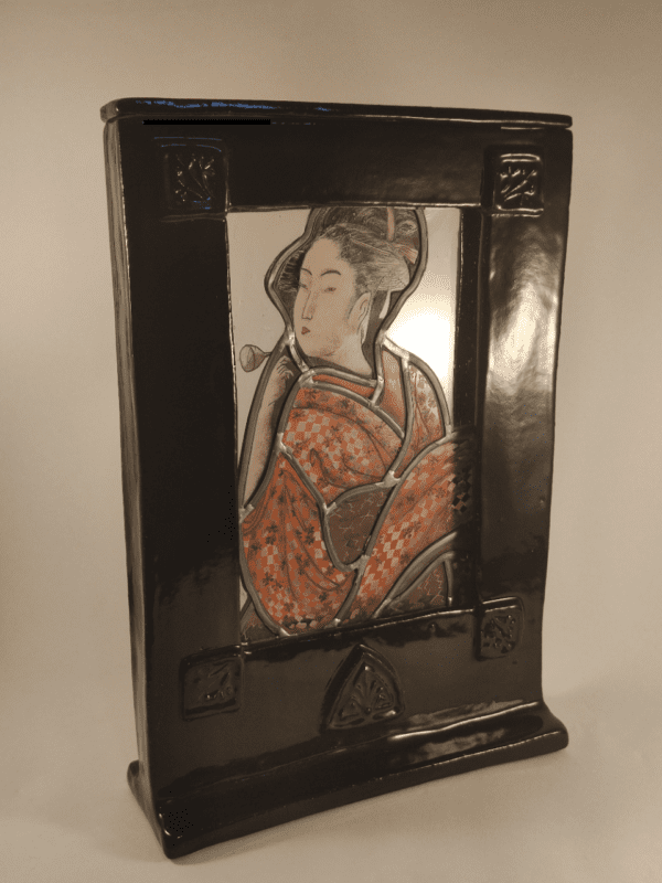 Cadre vitrail Terra. Un vitrail avec une geisha en kimono orange est inséré dans un cadre en céramique émaillé noire. Vue de trois quart